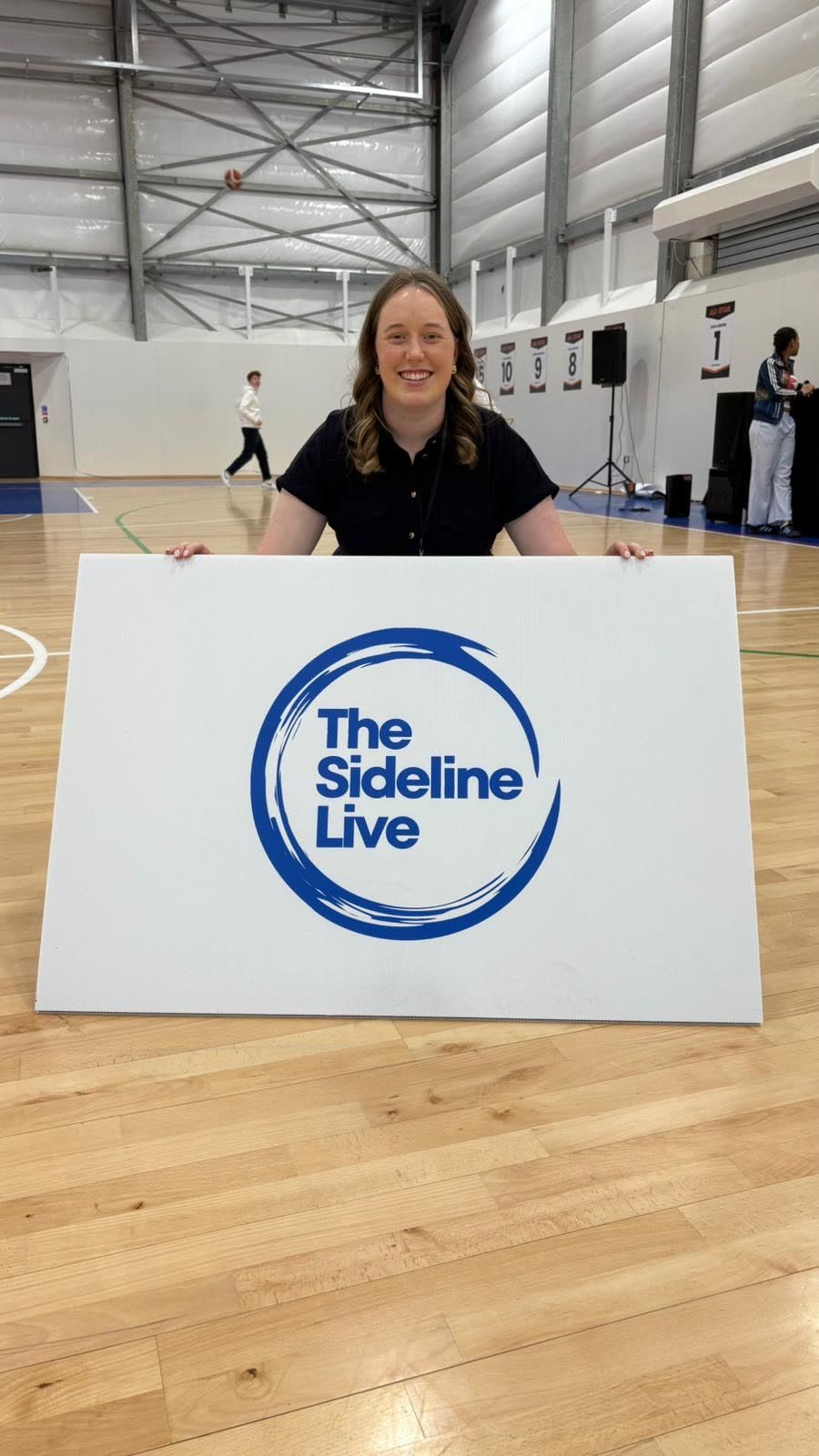 Orla Keeling on a basketball court, holding The Sideline Live sign