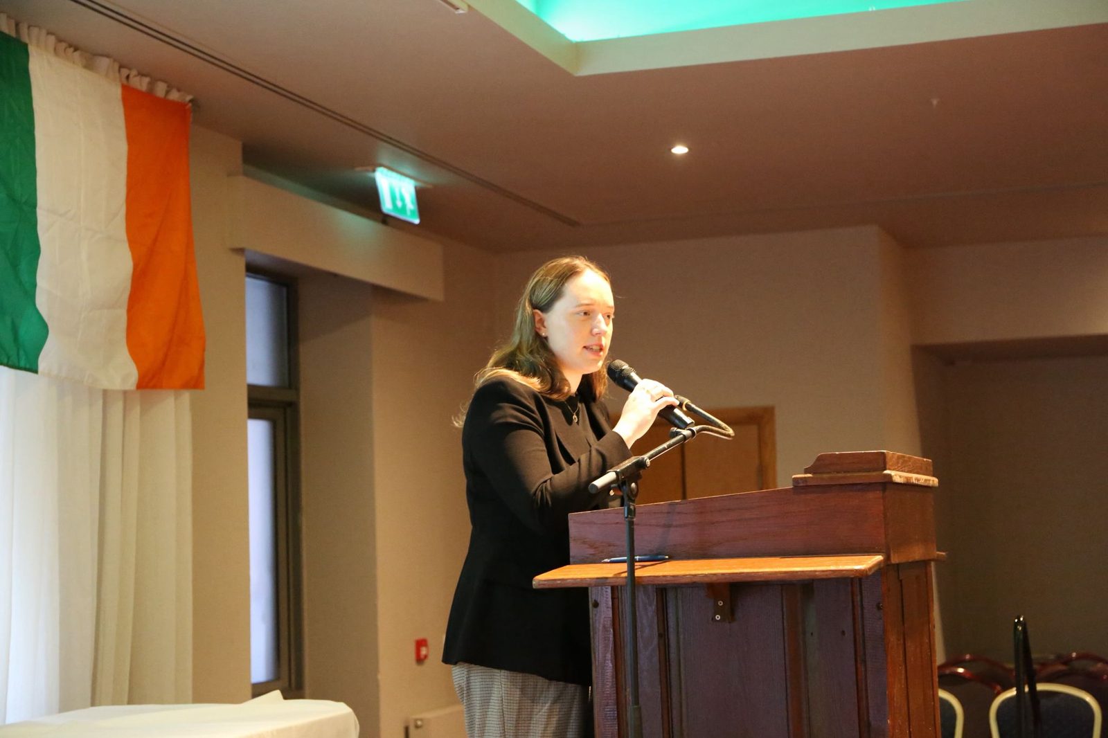Orla Keeling speaking at a lectern with a microphone, an Irish flag behind her